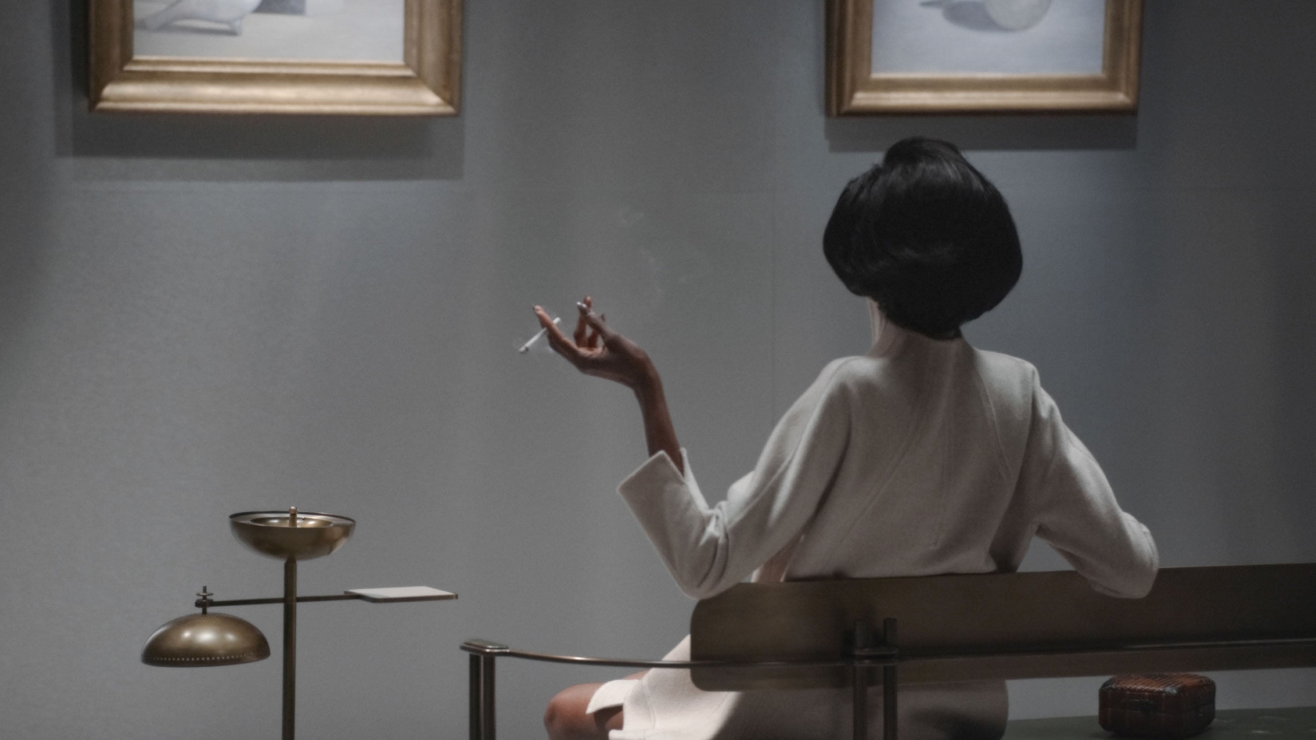 Woman in a white robe sitting on a bench with back turned, holding a cigarette in a dimly lit room with framed art.