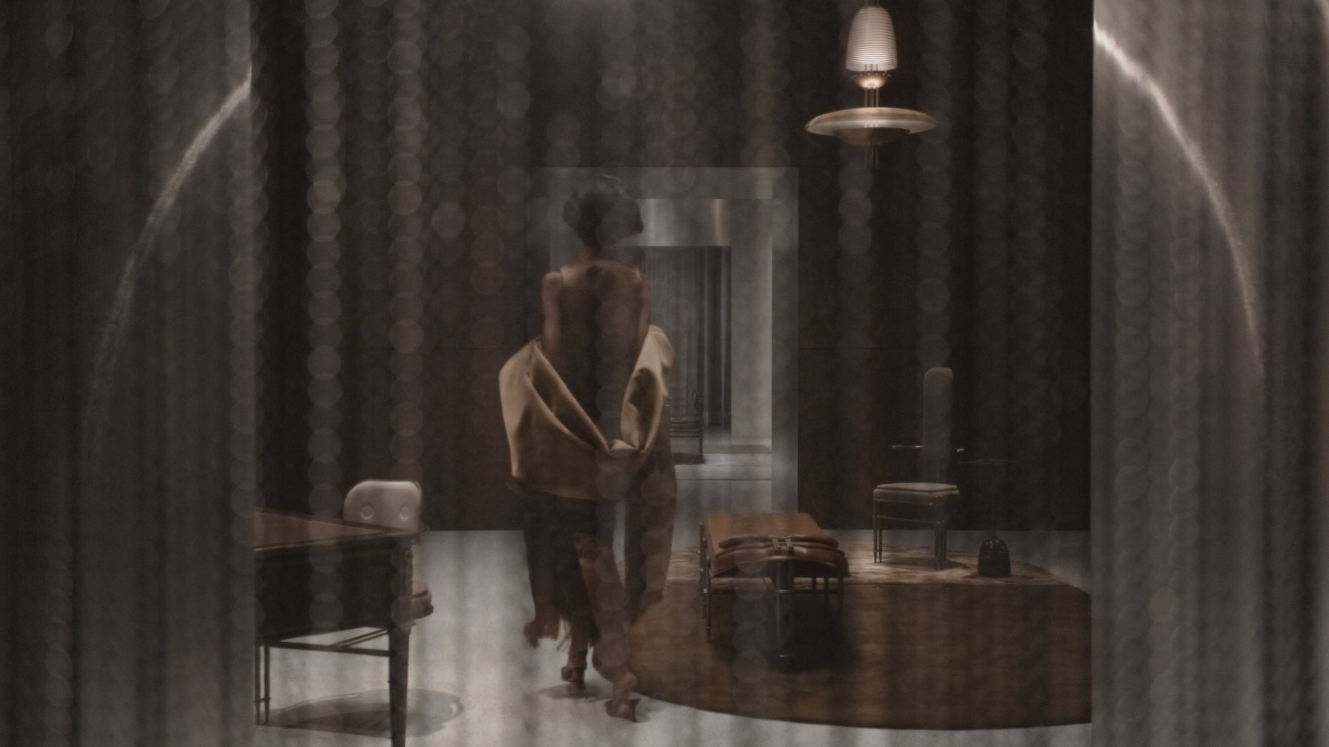 Dimly lit room with a person seated on a chair facing a mirror, surrounded by vintage furniture and soft lighting