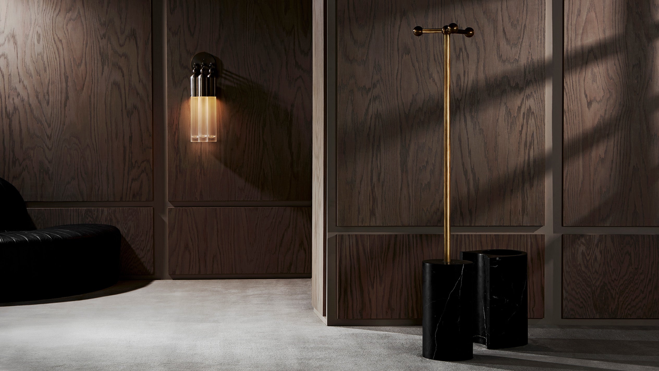 Modern interior with dark wood panel walls, black leather sofa, wall-mounted light fixture, and minimalist brass coat rack on concrete floor