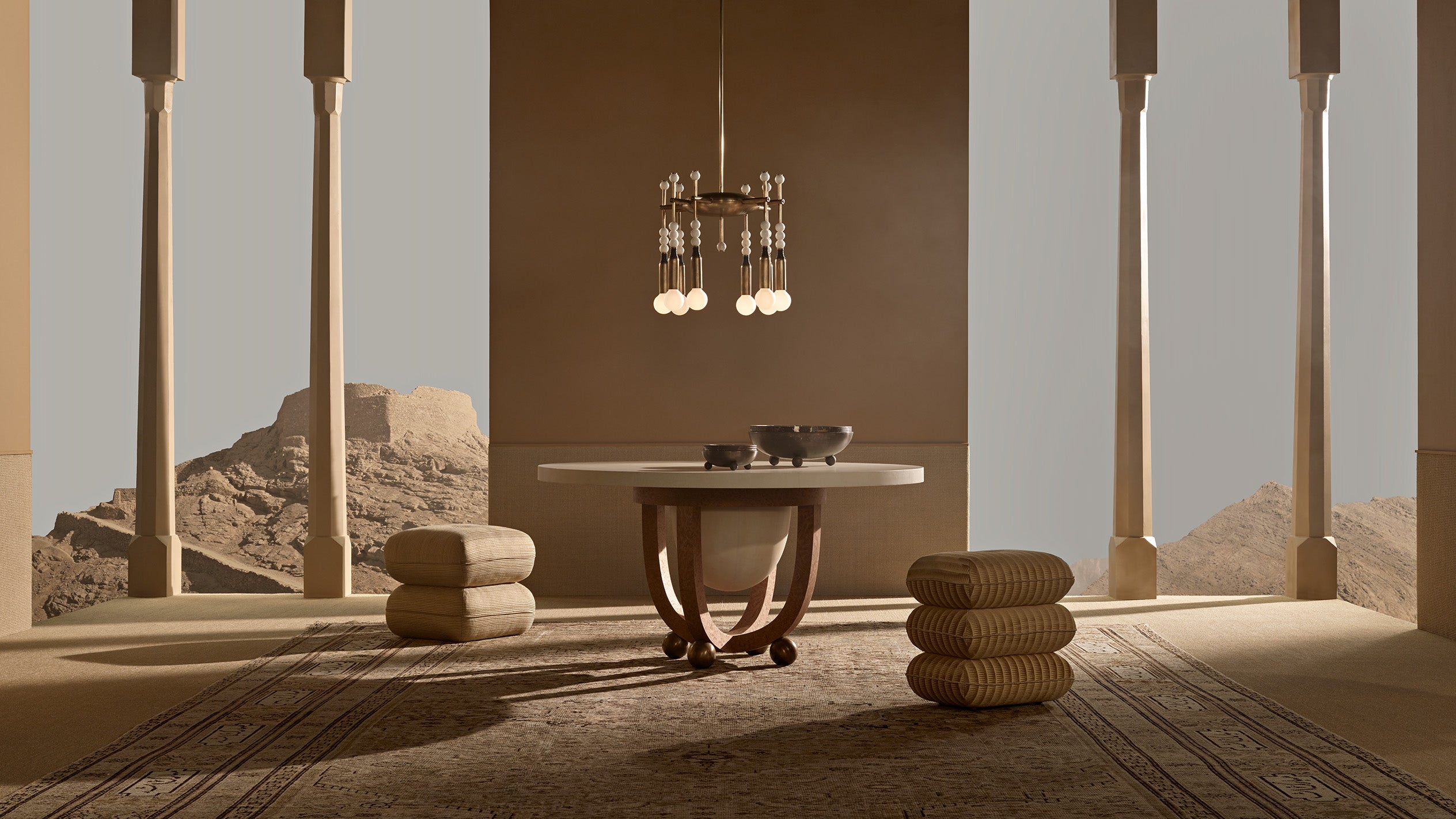 Minimalist interior with round stone table, stacked cushions, vertical columns, and a chandelier against an arid mountain backdrop