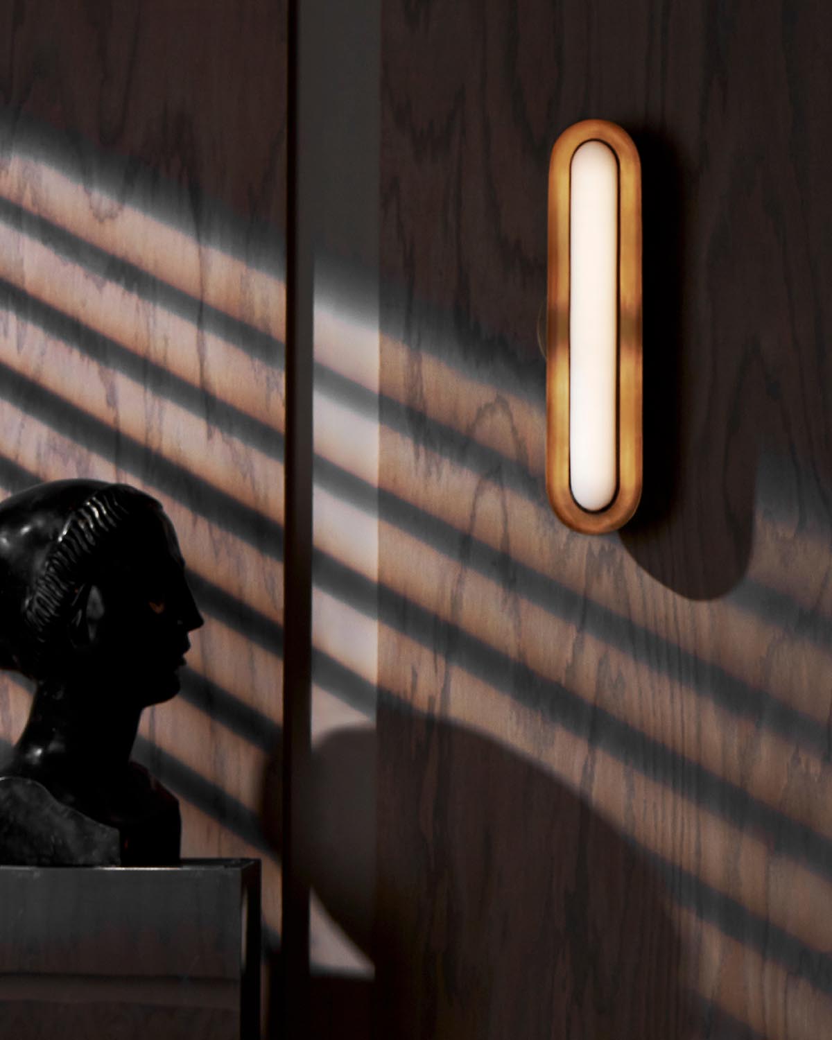 Wall-mounted oval light fixture with warm wooden frame casting shadows on dark wood paneling next to a dark bust sculpture