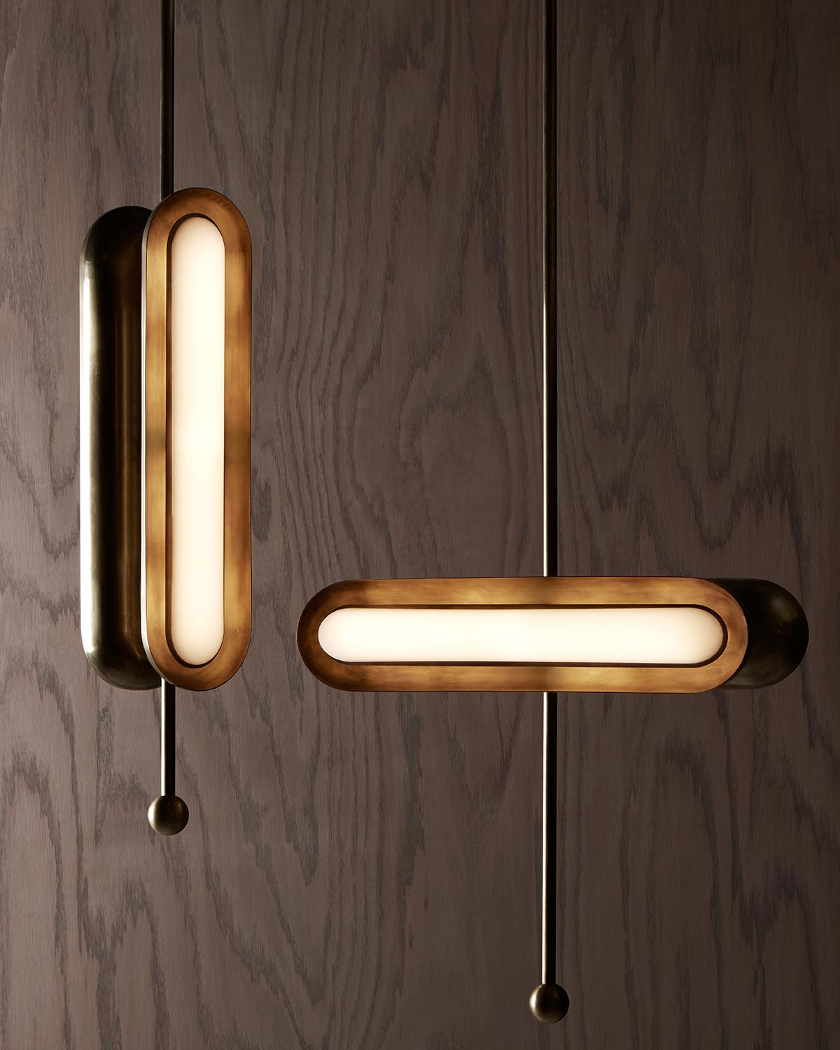 Two vertical pendant lights with elongated oval bronze frames and frosted glass diffusers against a wood panel background