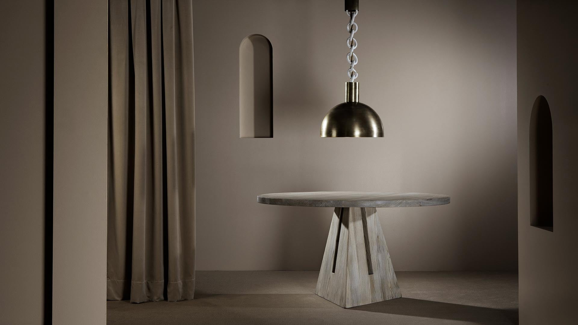 Round wooden table with geometric base under a brushed brass pendant light in a neutral-toned room