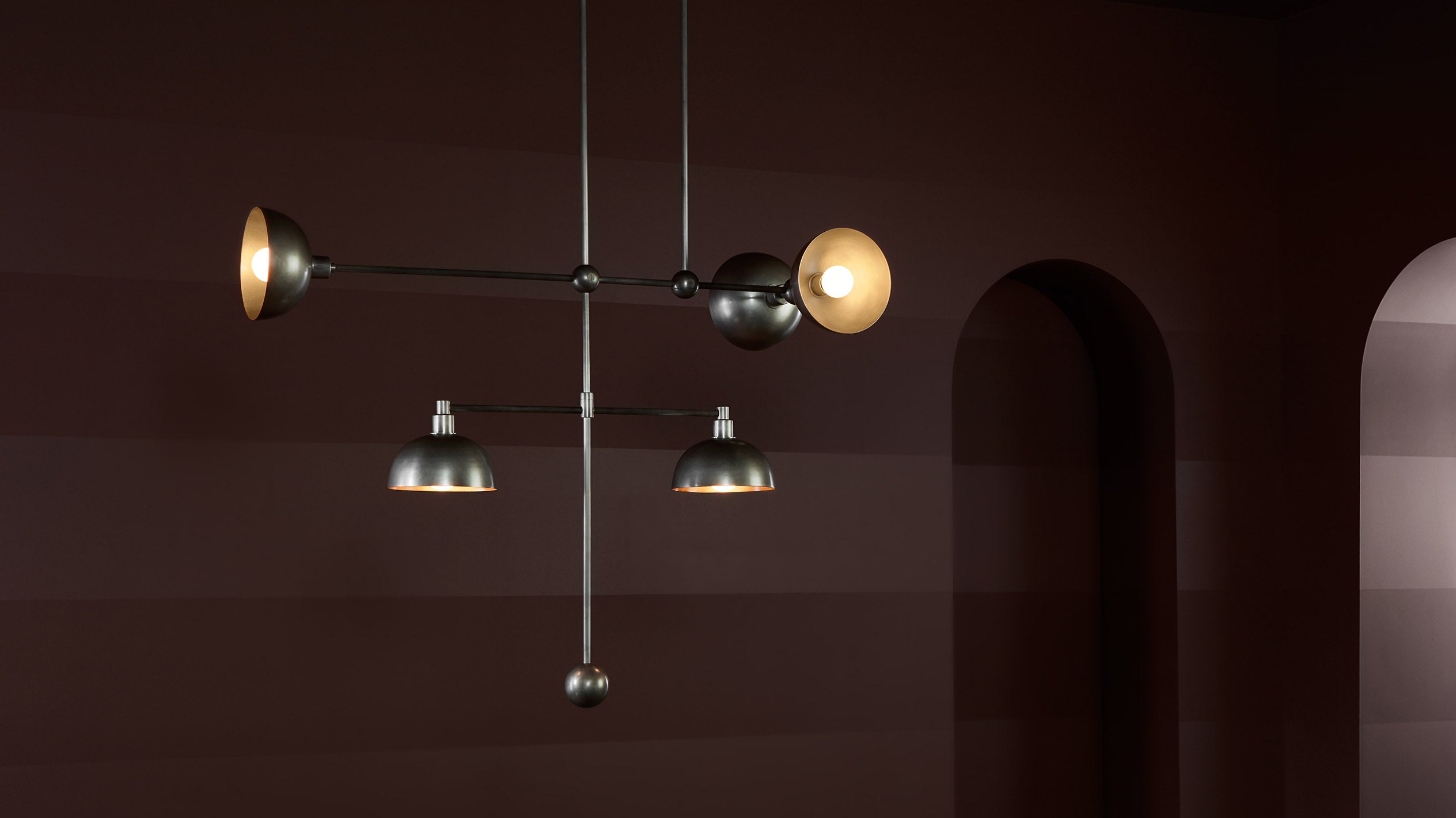 Modern black metal pendant light fixture with glowing bulbs hanging against a dark brown wall with arched doorways