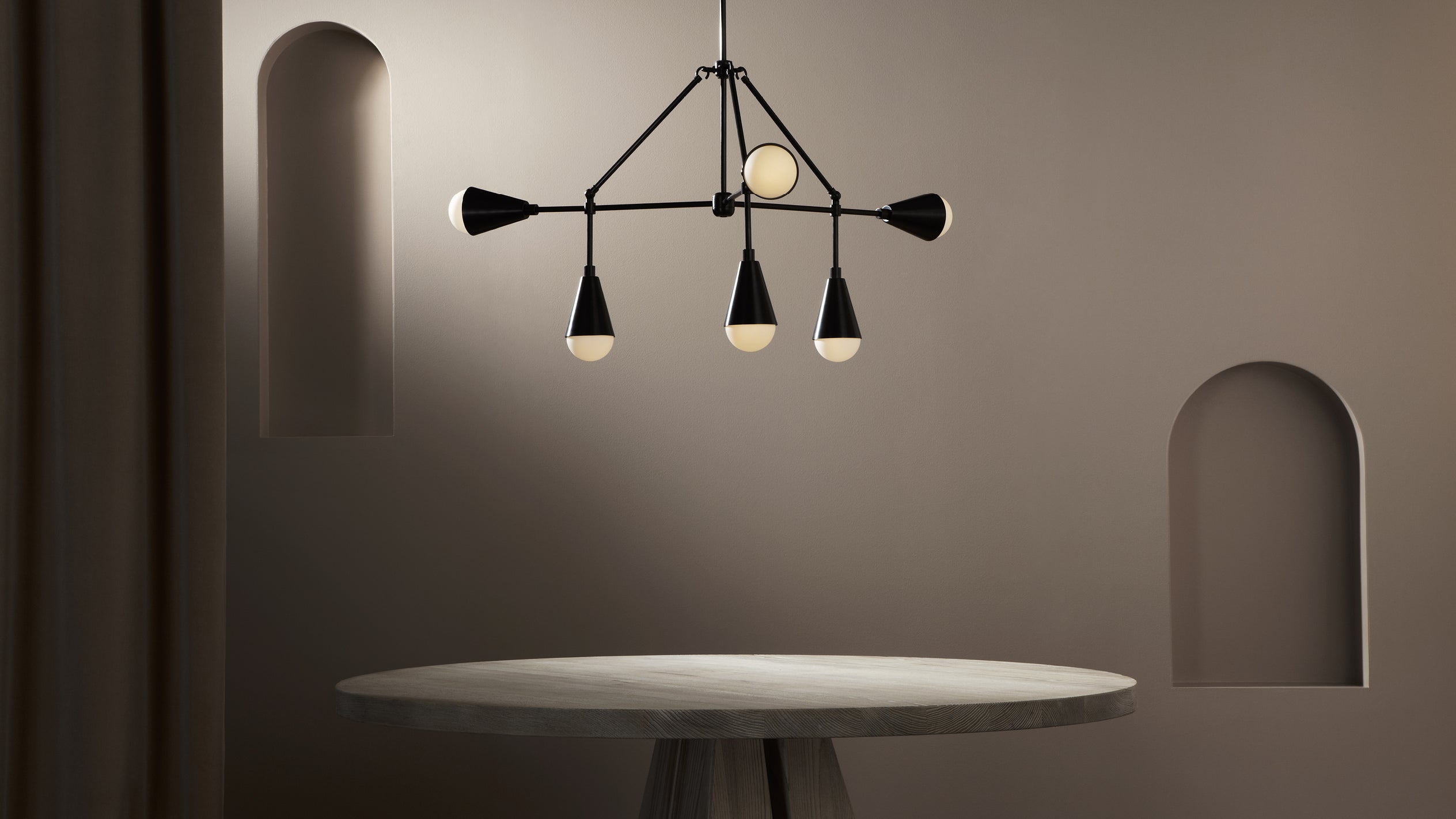 Round concrete table under a modern black metal chandelier with cone-shaped light bulbs in a beige minimalist room with arched wall niches