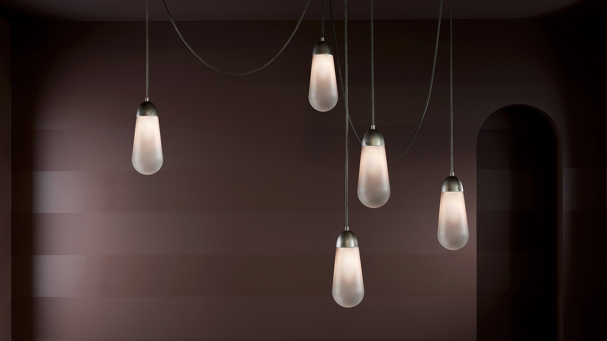 Five elongated pendant lights with frosted glass shades hanging against a dark brown textured wall with an arched opening