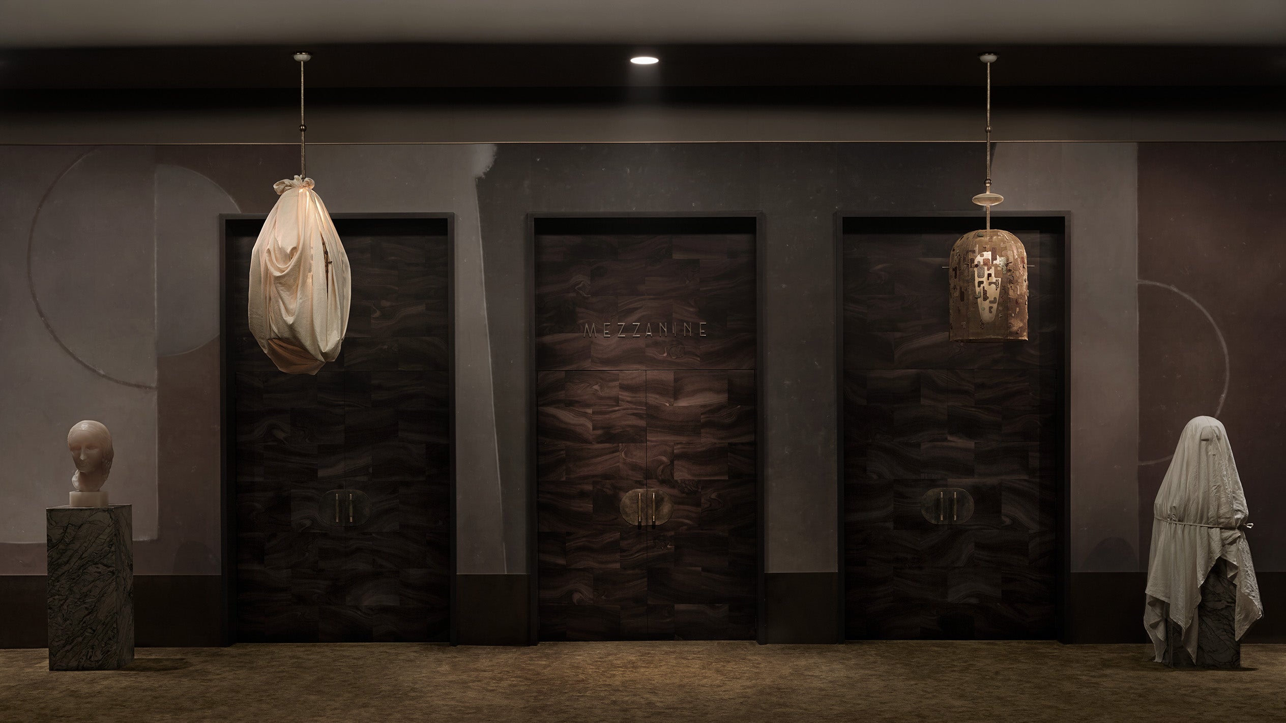 Dimly lit hallway with three ornate dark wooden doors flanked by two abstract sculptures and hanging pendant lights