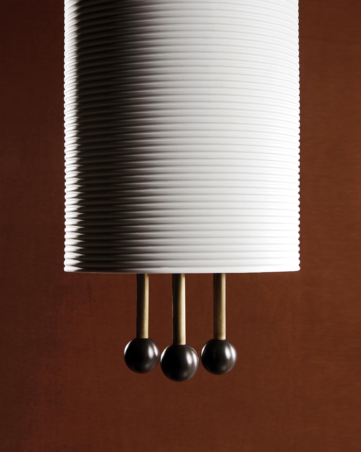 Close-up of white ribbed cylindrical pendant lamp with three brass rods ending in dark spherical accents against brown background