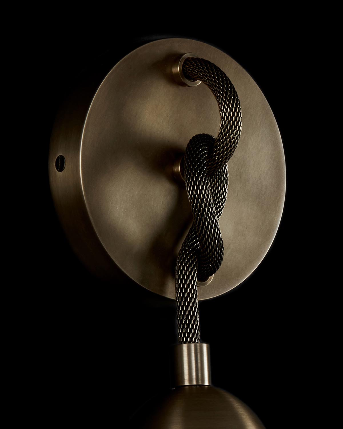 Close-up of a brass wall sconce with a black braided cord looped around the circular base against a black background
