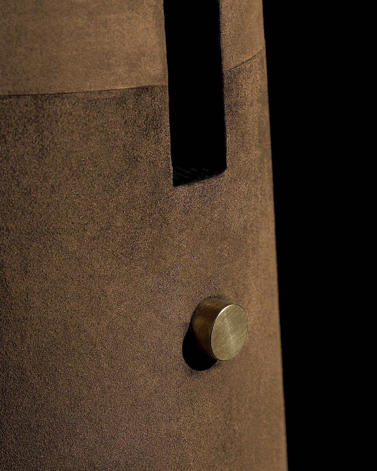 Close-up of brown suede texture with a small circular brass knob on the METRONOME table lamp body against black background