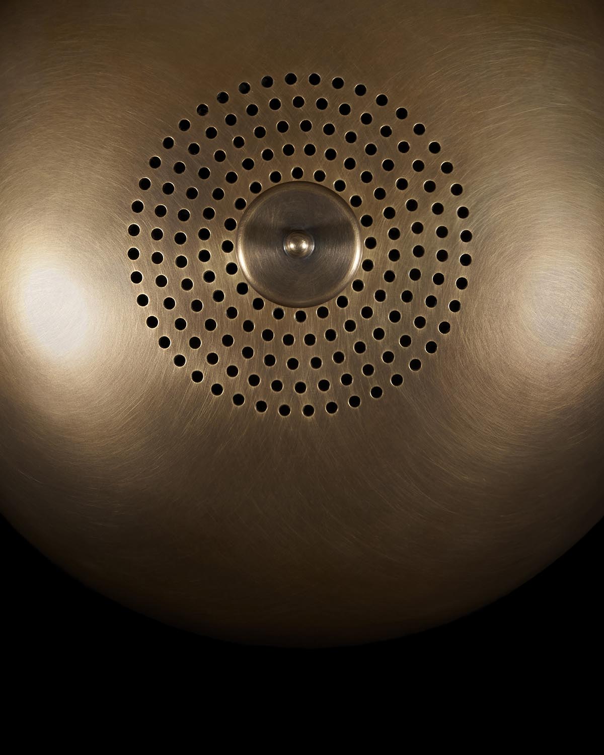 Top-down view of a circular metal vessel with concentric perforated holes and a central brushed knob on a dark background