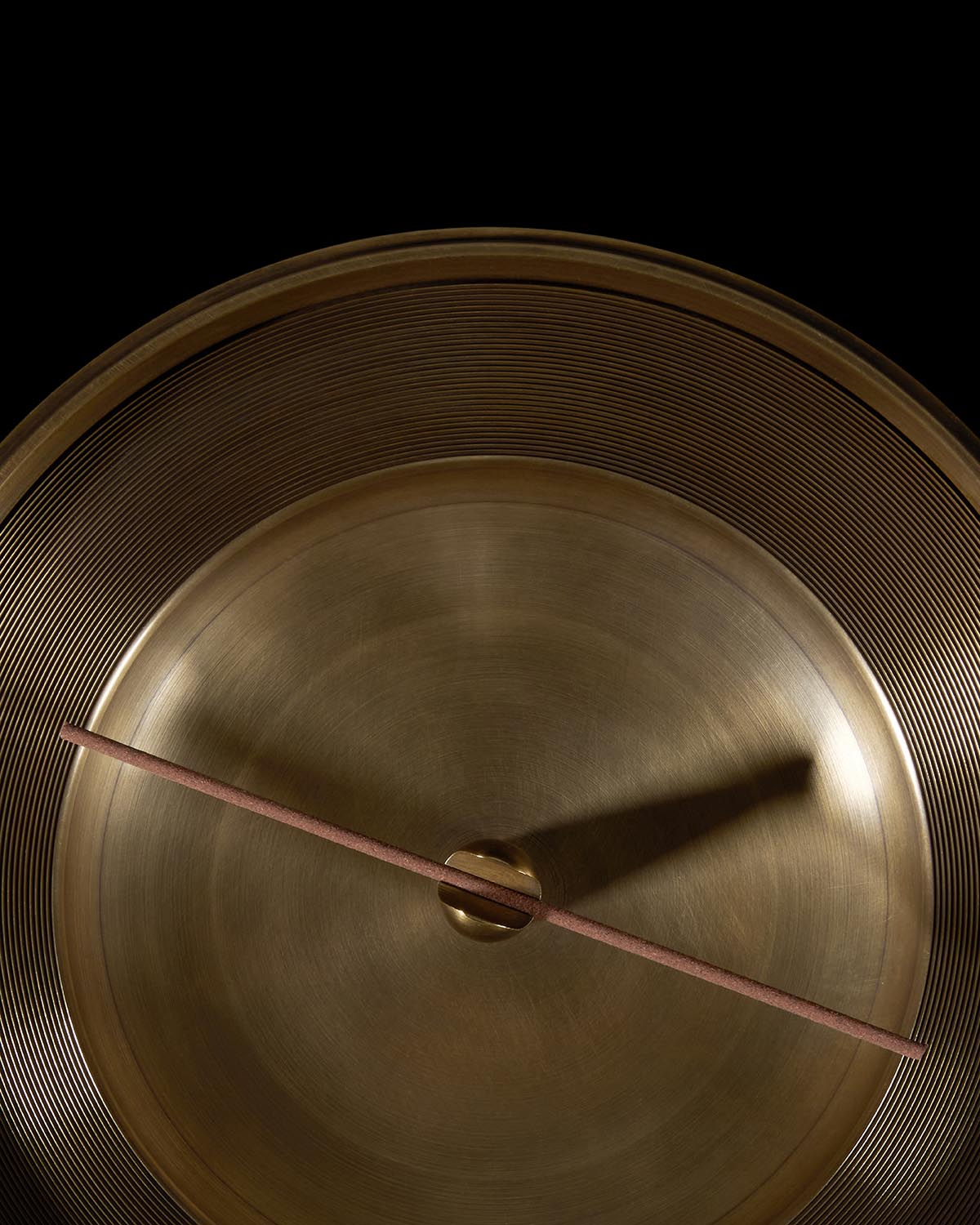 Top-down close-up of a brass smoking vessel with concentric ridges and a wooden stick resting across its center on black background