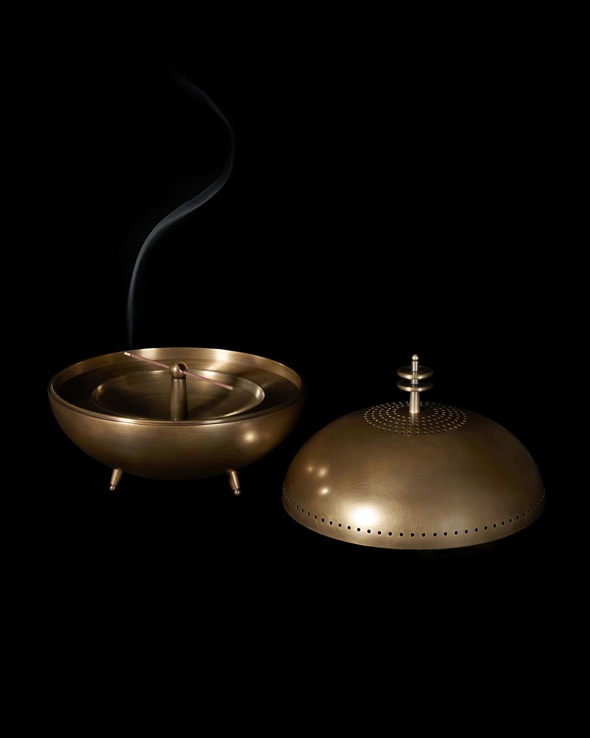 Brass smoking vessel with perforated dome lid and thin smoke rising against a black background