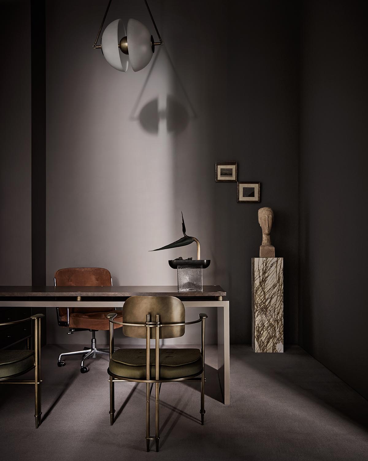 Minimalist office space with brown leather chair, olive green metal-framed chair, sculptural pendant light, and art on dark walls