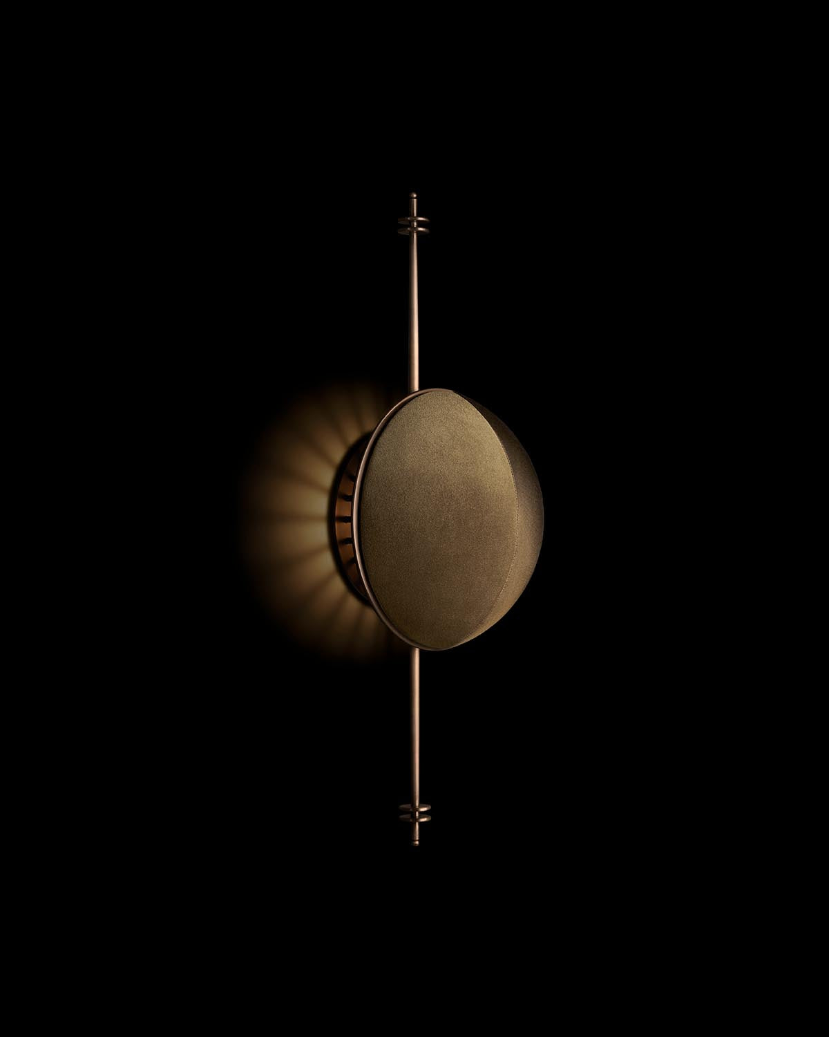 Vertical bronze wall light with circular shade casting a soft radial glow on black background