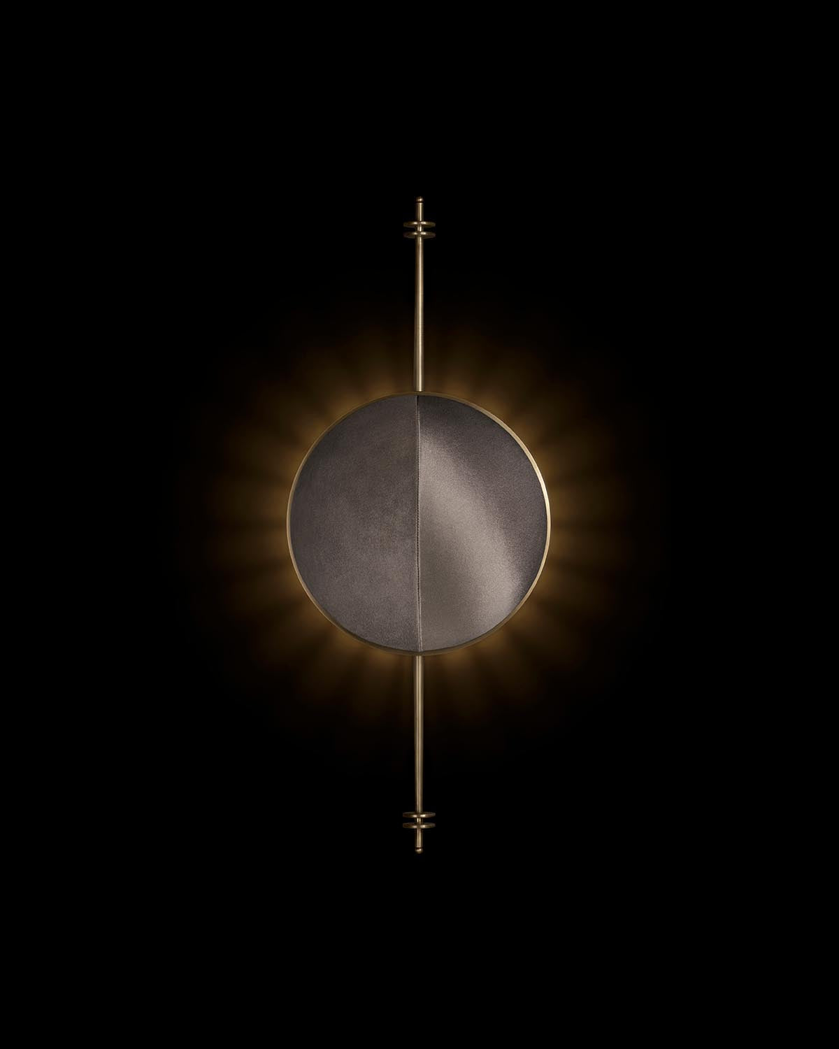 Minimalist round wall sconce with dark textured surface and vertical brass rod detail, glowing softly against black background