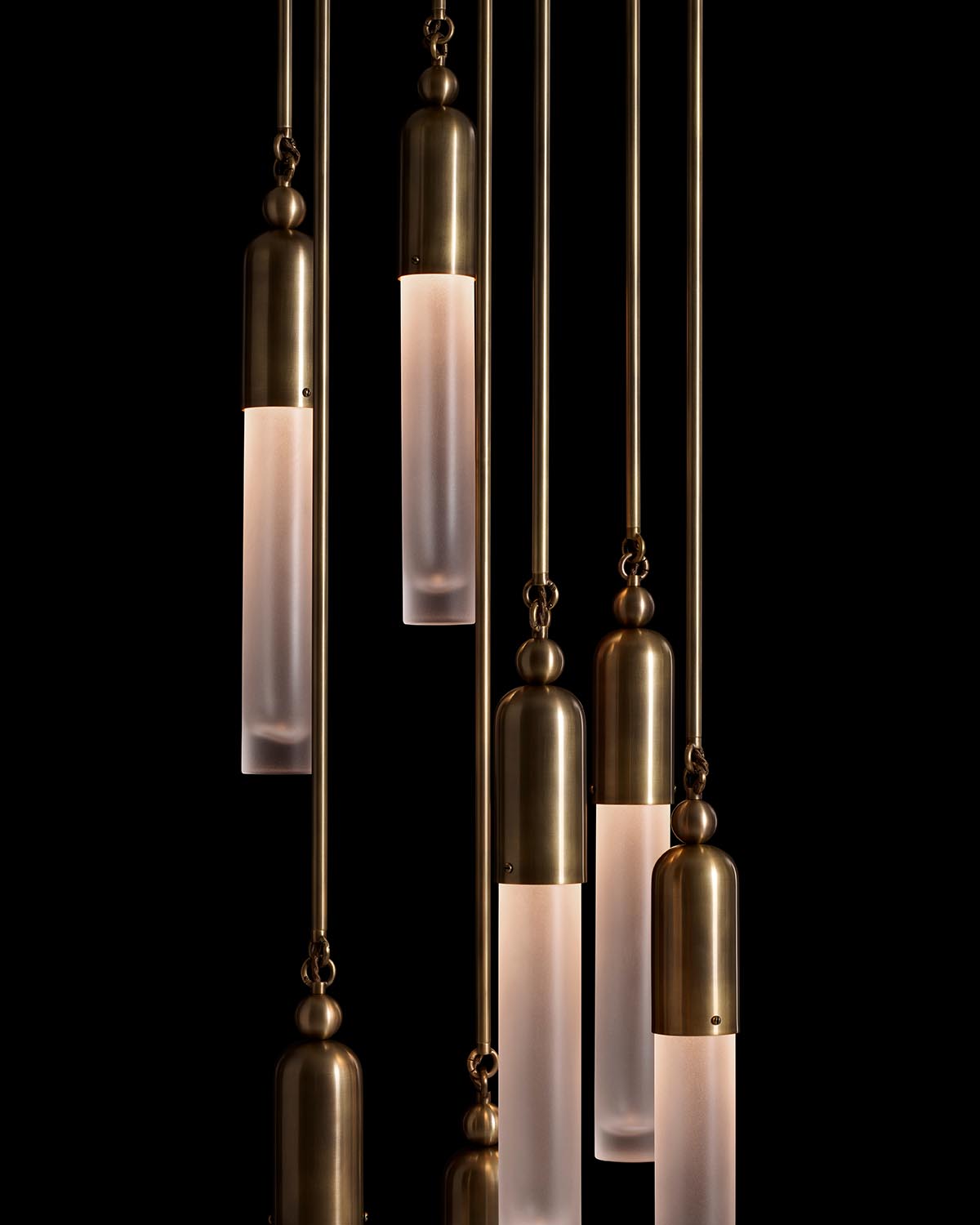 Seven vertical pendant lights with frosted glass tubes and antique brass fixtures hanging against black background