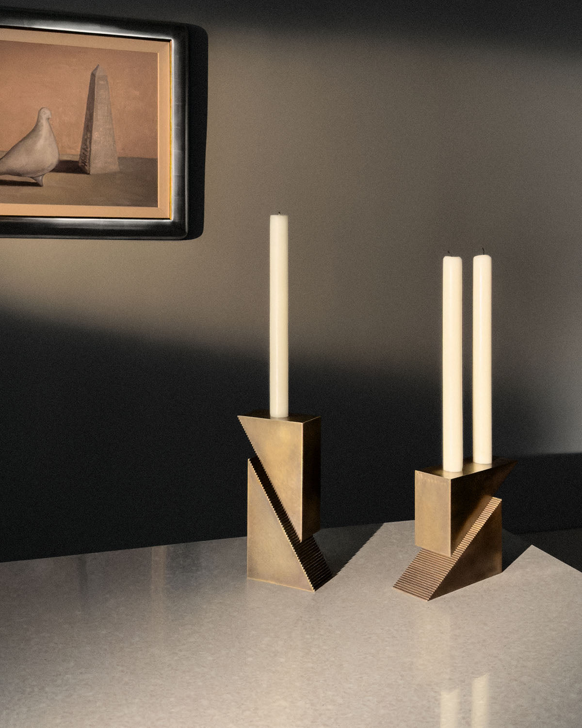 Pair of geometric brass candle holders each with one or two tall white taper candles on polished stone surface