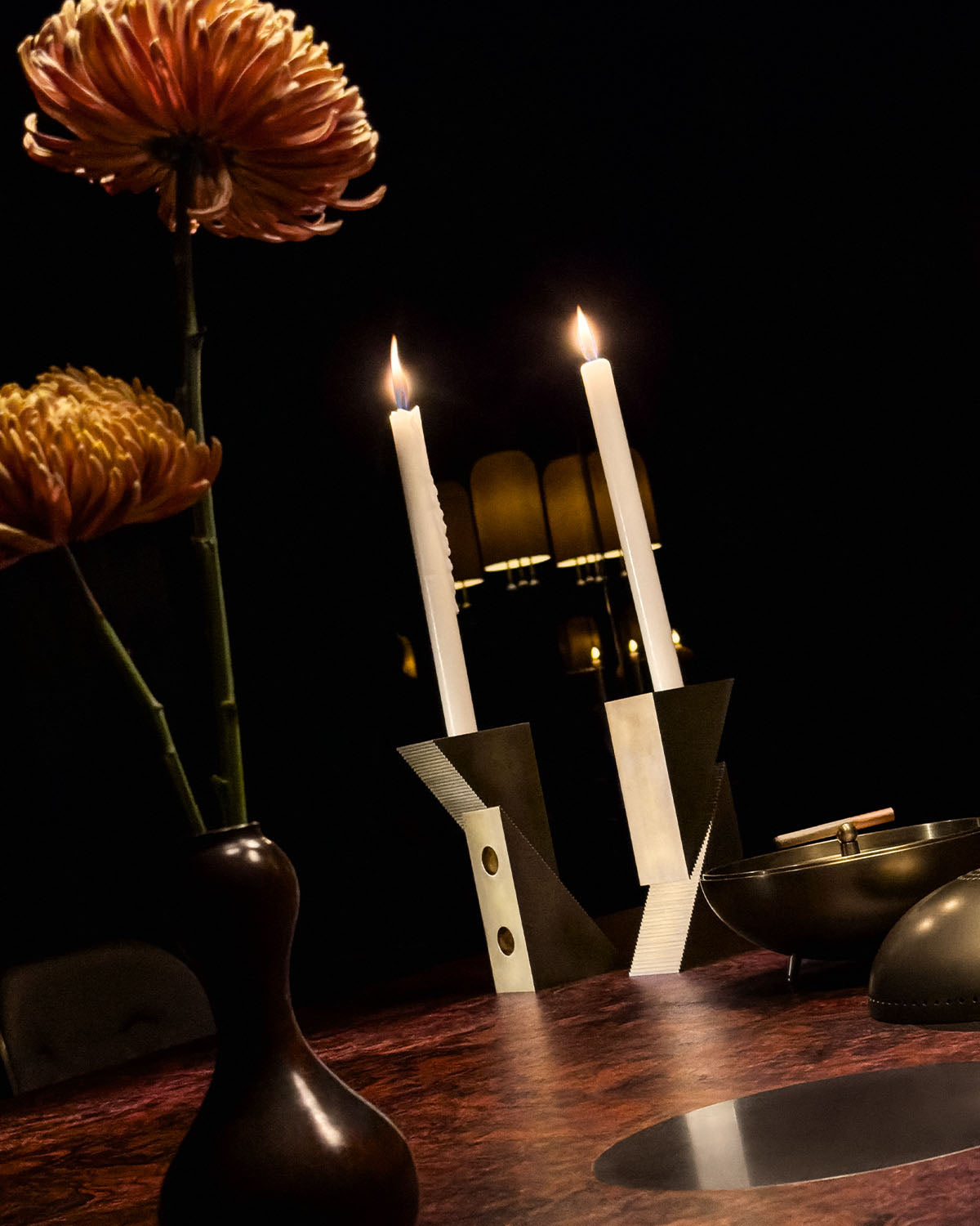 Two lit white taper candles in modern geometric candle holders on a dark wood table with flowers nearby